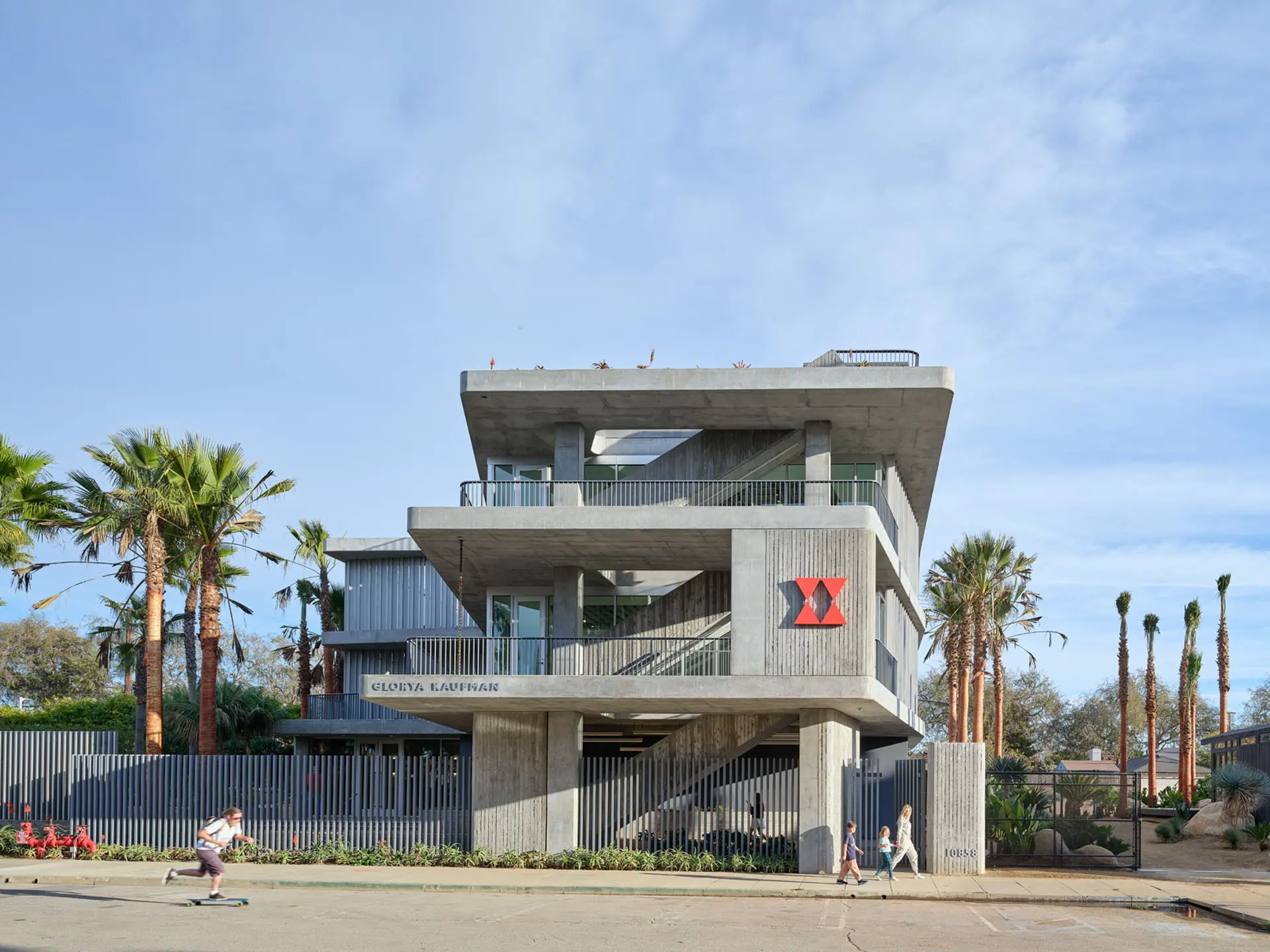View from road of three story concrete and glass building with palm trees out front and pedestrians and a skateboarder