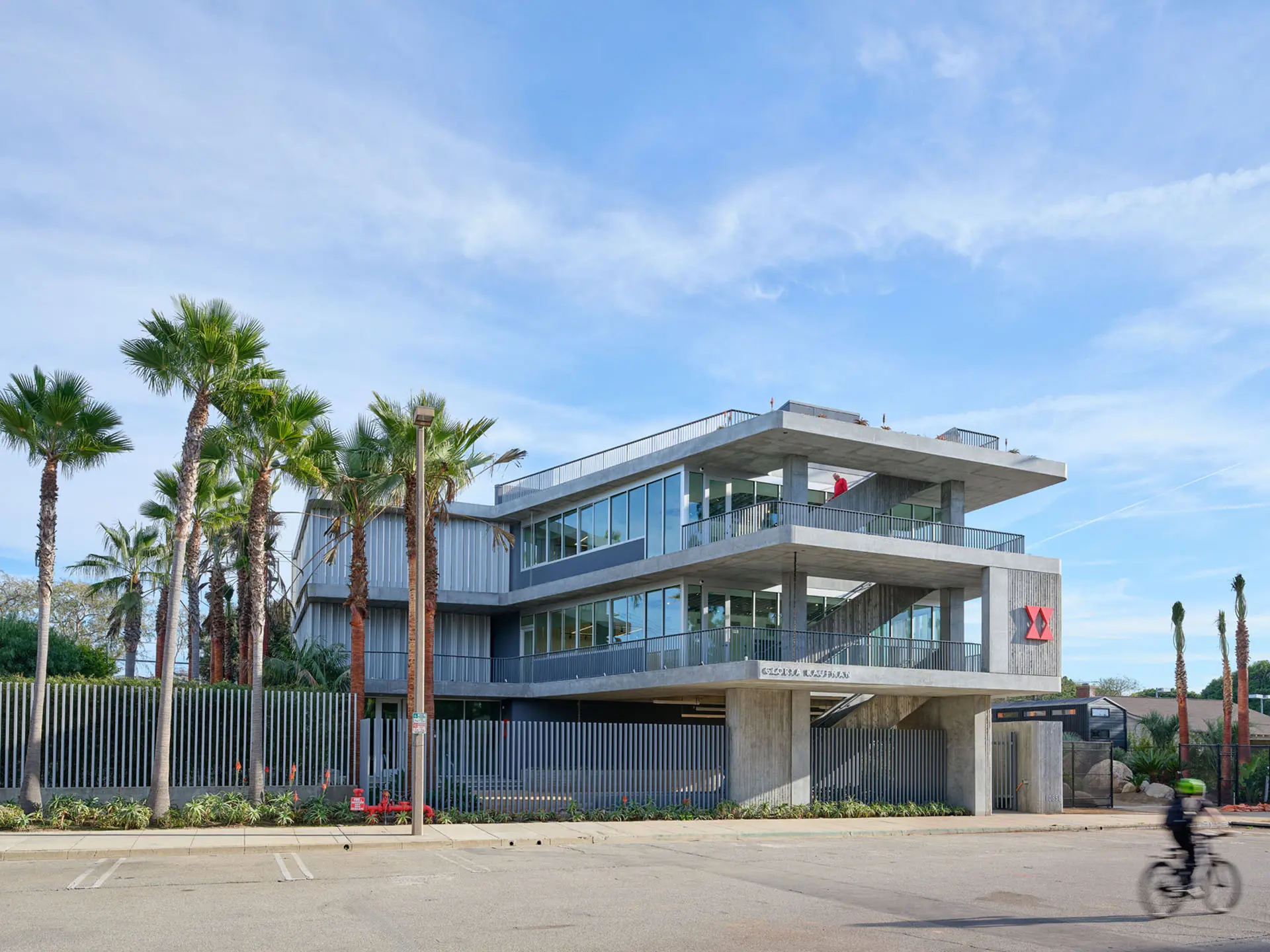 View from road of three story concrete and glass building with palm trees out front