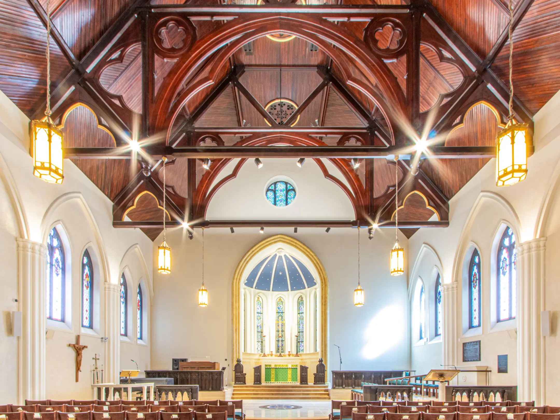 Interior image of wooden ceiling of church sanctuary with tabernacle, stained glass windows, and pendant lights